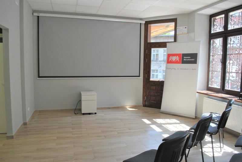 zdjęcie przedstawia wyremontowane pomieszczenie na potrzeby Stowarzyszenia WAGA i Centrum Wsparcia Seniora, służące do spotkań. Na ścianie umieszczony został ekran projekcyjny.