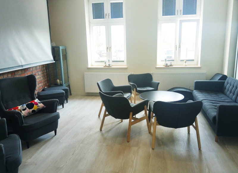 Zdjęcie przedstawia wyremontowane pomieszczenie spełniające funkcję głównego miejsca spotkań z przeznaczeniem na potrzeby fundacji FOSA i Centrum Wsparcia i Terapii Rodzin. W pomieszczeniu ekran projekcyjny, czarna sofa i fotele.