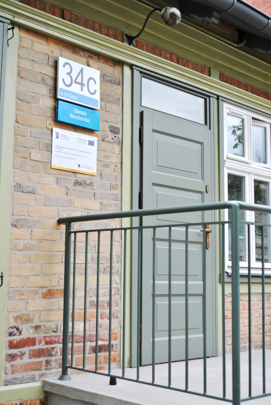 zdjęcie przedstawia wyremontowane wejście do budynku. Drzwi wejściowe z elementem poręczy i tablicami informacyjnymi.