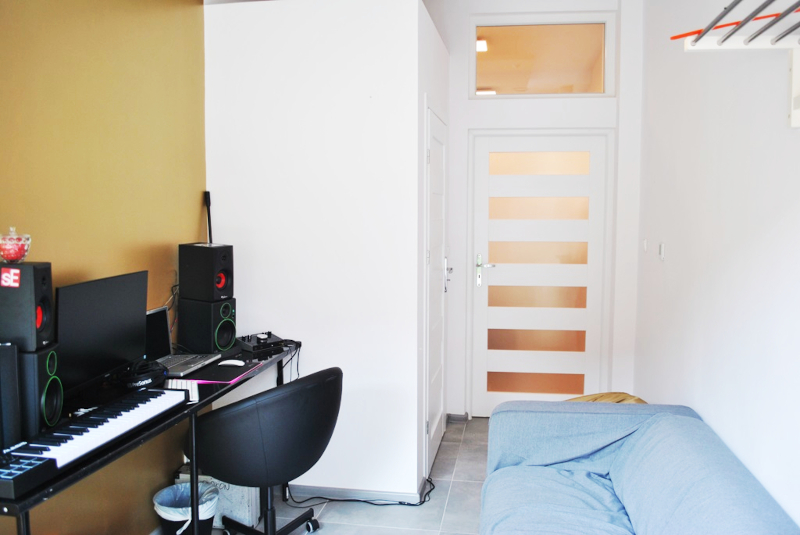 zdjęcie przedstawia wyremontowany pomieszczenie. Po lewej stronie znajduje się biurko ze sprzętem muzycznym a po prawej szara sofa.