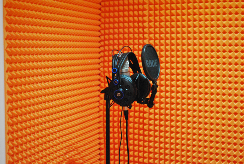 Na pierwszym planie znajduje się profesjonalny mikrofon umieszczony w wyremontowanym studio w intensywnym pomarańczowym kolorze.