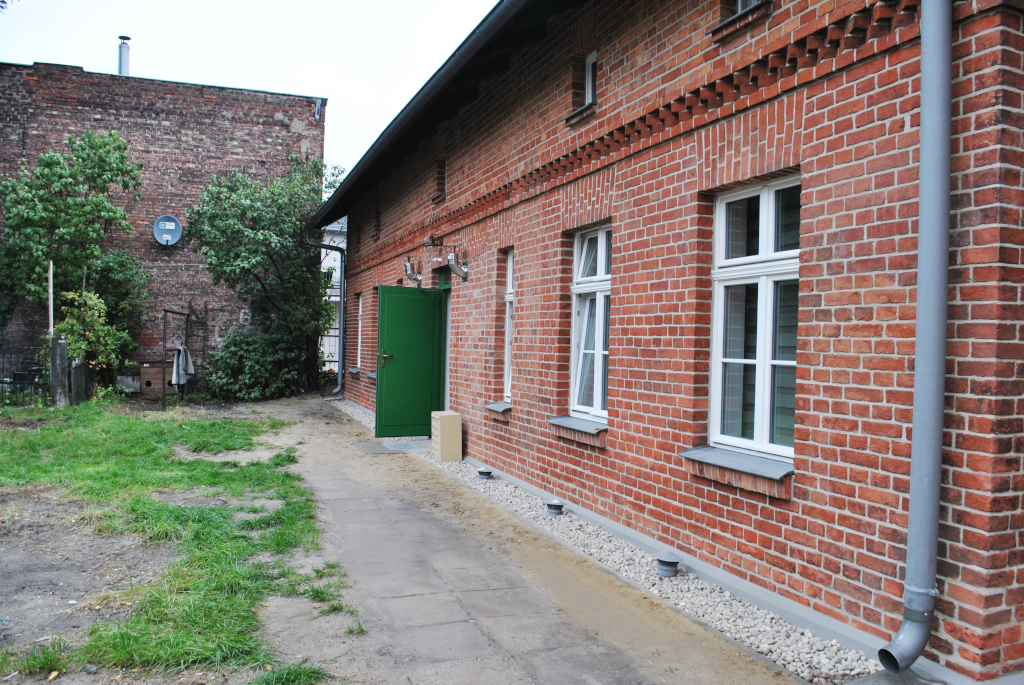 zdjęcie przedstawia budynek po rewitalizacji od strony podwórza z uchylonymi zielonymi drzwiami, które prowadzą do budynku.
                 Na drugim planie ściana innego budynku z czerwonej cegły.